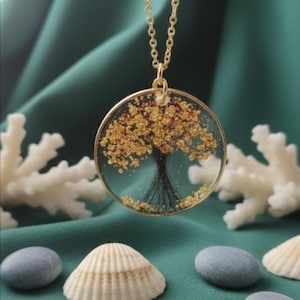Puede incluir: Un collar dorado con un colgante circular con un diseño de árbol. El árbol tiene un tronco negro y hojas doradas. El colgante cuelga de una delicada cadena dorada. El fondo incluye conchas marinas, piedras y coral.