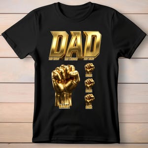 以下が含まれることがあります： 黒の半袖Tシャツ。金色の文字で「DAD」「THE HERO」「THE LEADER」「THE KING」とプリントされている。その下には大きな金色の拳のグラフィックがあり、その下に「NAME」という文字がある。さらにその横に、それぞれ「NAME」と書かれた小さな金色の拳のグラフィックが3つ並んでいる。
