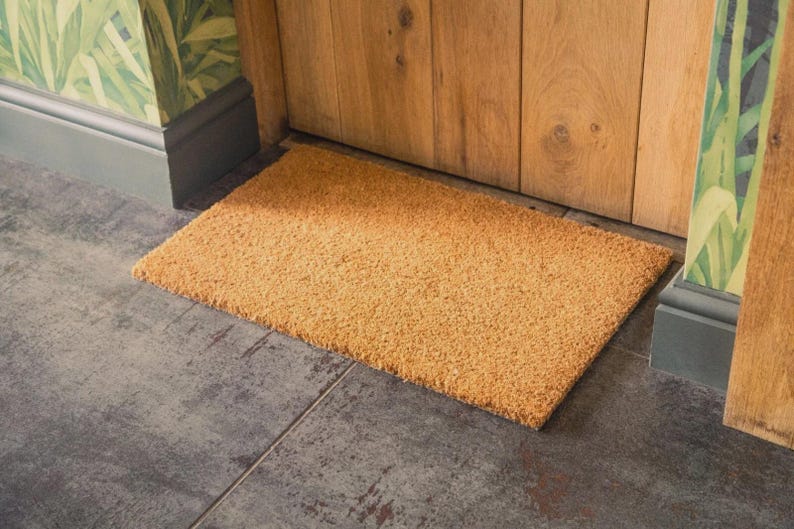 Pu&ograve; includere: Un zerbino rettangolare in fibra naturale, di colore marrone chiaro, &egrave; posizionato su un pavimento piastrellato. Il tappeto &egrave; posto davanti a una porta di legno, con carta da parati a motivi visibile sulla parete.