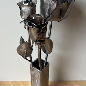 Può includere: Una scultura in metallo di tre rose in un vaso quadrato. Le rose sono realizzate in metallo color argento, con petali e foglie dettagliati. Il vaso è anch'esso in metallo, creando un'esposizione artistica. È un oggetto decorativo.