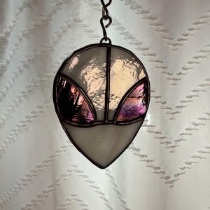 May include: A stained glass alien head suncatcher. The alien head is primarily white with iridescent accents. The eyes are a deep purple. The piece is framed with a dark metal and hangs from a metal chain.