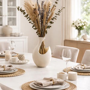 Peut inclure: Mise en place pour un dîner sur une table ovale blanche. Les couverts comprennent des serviettes beiges, des ronds de serviette dorés et des sets de table tressés. Un vase blanc contient des fleurs séchées.