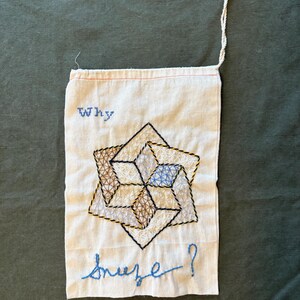 May include: Embroidered fabric art with the words "Why sneeze?" in blue thread. The artwork features a geometric design in black, brown, yellow, and blue thread. A white cord is attached to the top.