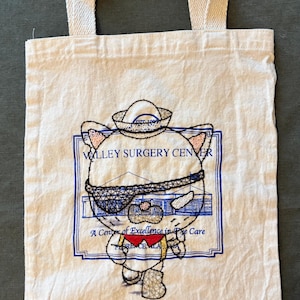 Puede incluir: Bolso tote de lona color crema con asas color crema. El bolso presenta un diseño bordado de un gato con gorro de marinero y gafas de sol, con el texto "Valley Surgery Center" y "A Center of Excellence in Care".