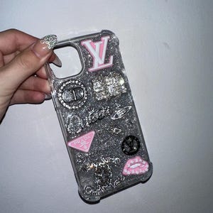 May include: A clear phone case filled with silver glitter, decorated with various charms including a pink LV logo, a crystal CD emblem, a crystal handbag, a pink Prada triangle logo, a black circular logo, and a pink lip-shaped charm.