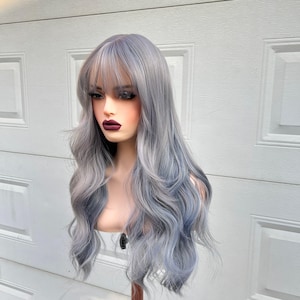 Puede incluir: Una peluca larga y ondulada en tonos grises y azules, con flequillo. La peluca se muestra en una cabeza de maniquí, mostrando su diseño en capas y variaciones de color. El cabello tiene una textura de aspecto natural y es adecuado para cosplay o uso diario.