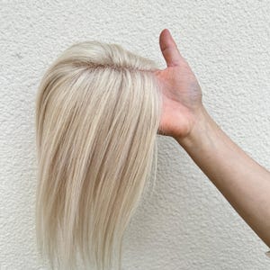 May include: A light blonde hair topper with long, straight strands. The hairpiece is held up, showcasing the natural-looking roots and the overall color. The hairpiece appears to be made of human hair, and is designed to add volume and coverage.