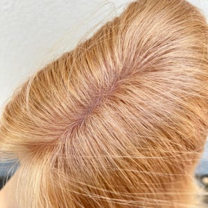 May include: Close-up of a blonde wig's lace front and hair strands. The hair is a light, warm blonde color, with a natural-looking hairline. The lace is a light brown color, designed to blend with the skin.