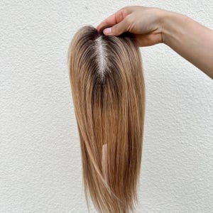 以下が含まれることがあります： 白い背景に、暗い根元を持つ明るい茶色のヘアトッパーが保持されています。髪はストレートで、長さは約30cmです。トッパーは自然な見た目の分け目があります。