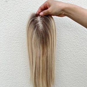 Op de afbeelding: Een haarstuk met steil, meerkleurig blond haar en donkerbruine wortels. Het item heeft een zichtbare monofilament basis bij de kruin, wat een realistische hoofdhuid laat zien waar de haarlokken individueel zijn ingeknoopt.