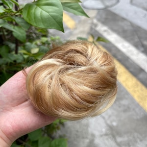 May include: A synthetic hair bun extension featuring a blend of light blonde and golden blonde highlights. The hairpiece is styled in a circular, voluminous shape with a textured, layered appearance, designed to be worn as a hair accessory.