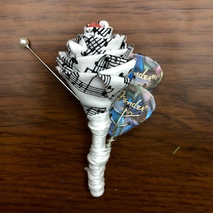 May include: A unique boutonniere featuring a fabric rose with musical notes, accented with Fender guitar picks. The rose is white with black musical notes, and the stem is wrapped in white thread. A pearl-headed pin is included.