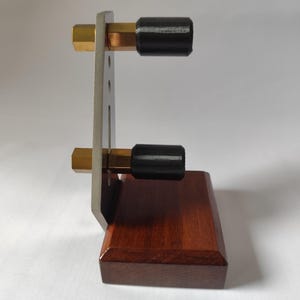 May include: A wooden and metal object with a rectangular base. The base is dark brown. A silver metal plate is attached to the base. Two brass-colored hexagonal bolts extend from the plate, each with a black cylindrical rubber end. The object is on a white surface.