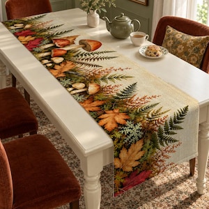 Puede incluir: Un camino de mesa rectangular con un estampado de temática otoñal que presenta setas naranjas y rojas, hojas de otoño en tonos dorados, naranjas y carmesíes, y helechos verdes sobre una tela de textura beige similar al lino.