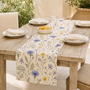 May include: A rectangular table runner featuring a botanical print of blue cornflowers, yellow buttercups, and green foliage on an off-white background. The runner is centered on a wooden table with white plates, napkins, and clear glasses.