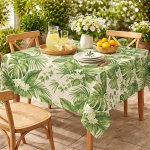 May include: A rectangular tablecloth with a tropical leaf pattern in shades of green and white covers a wooden table. The table is set with plates, cutlery, and a bowl of lemons. Two wooden chairs are visible.