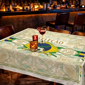 May include: A white tablecloth with a green and gold geometric design, featuring the words "Canarinho Spirit" and "The Land of Samba." The tablecloth is decorated with a Brazilian flag and a soccer ball. A cocktail, a candle, and a glass of whiskey are on the table.