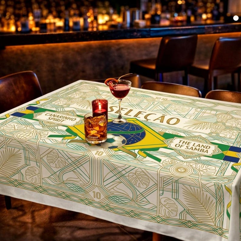 May include: A white tablecloth with a green and gold geometric design, featuring the words "Canarinho Spirit" and "The Land of Samba." The tablecloth is decorated with a Brazilian flag and a soccer ball. A cocktail, a candle, and a glass of whiskey are on the table.