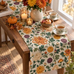 Peut inclure: Un chemin de table couleur crème orné d'un motif botanique répétitif de chrysanthèmes orange, jaunes et violets avec des feuilles vertes. Le chemin de table est présenté sur une table en bois avec un pichet en céramique, des bougies et des tasses.
