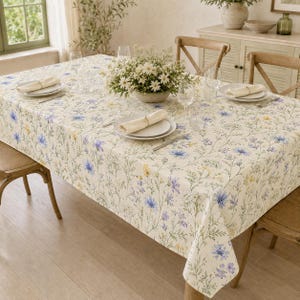 Puede incluir: Un mantel rectangular con fondo crema y un delicado estampado botánico de flores silvestres azules y amarillas. La mesa está puesta con platos blancos, servilletas crema dobladas, cristalería transparente y un florero central.