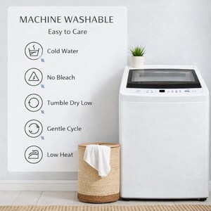 May include: A white top-load washing machine with a black control panel. A woven laundry basket with a white towel is in front of the machine. Text on a sign indicates the machine is machine washable and easy to care for.