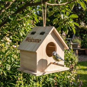 Puede incluir: Una pajarera de madera clara con un tejado inclinado y un agujero de entrada redondo. La palabra "Welcome" está grabada en la parte delantera. Un pequeño pájaro azul y amarillo se posa en un palito de madera. La pajarera cuelga de una cuerda.
