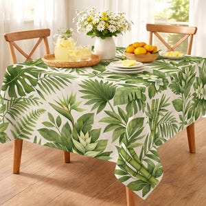 May include: A rectangular tablecloth with a tropical leaf pattern in shades of green on a cream background. The table is set with a pitcher of lemonade, glasses, and a bowl of lemons. The table is surrounded by wooden chairs.