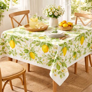 Op de afbeelding: Een witte tafelkleed met een citroen- en bloemenpatroon bedekt een houten tafel. De tafel is gedekt met een limonadekan, glazen, borden en een kom met citroenen. Een vaas met madeliefjes staat in het midden.