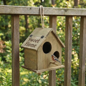 Puede incluir: Una pajarera de madera con el texto "Sweet Home" en la parte delantera. La pajarera tiene dos aberturas circulares y una pequeña percha. Un pequeño pájaro con plumas marrones, naranjas y grises está posado en la pajarera. La pajarera está colgada de una valla de madera.