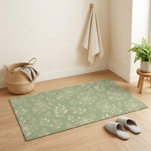 Könnte beinhalten: Ein rechteckiger, salbeigrüner Teppich mit einem floralen Muster aus weißen Blättern und Blüten. Der Teppich liegt auf einem Holzboden, mit einem Paar grauer Hausschuhe in der Nähe. Ein geflochtener Korb und ein Handtuch hängen ebenfalls im Raum.