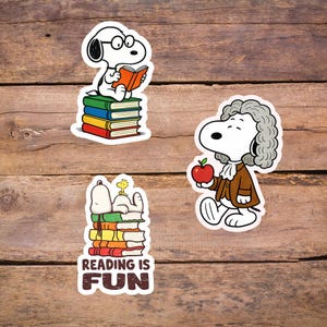 Könnte beinhalten: Set mit drei Snoopy-Aufklebern. Ein Aufkleber zeigt Snoopy, der ein Buch auf einem Stapel von Büchern liest. Ein anderer zeigt Snoopy als Isaac Newton mit einem Apfel. Der dritte Aufkleber zeigt Snoopy und Woodstock mit dem Text "READING IS FUN".