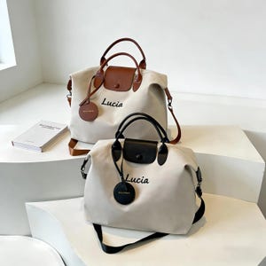 Pode incluir: Duas malas tote creme com detalhes em castanho e preto. As malas têm alças superiores e alças de ombro. A palavra "Lucia" está escrita em letras pretas na frente de cada mala. Uma mala tem uma aba castanha e a outra uma aba preta.