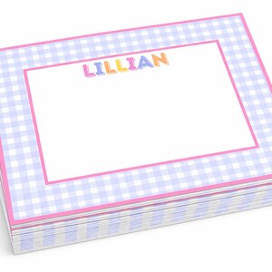 Puede incluir: Una pila de blocs de notas personalizados. Cada bloc presenta un interior blanco con un borde rosa y un patrón de cuadros vichy azul claro y blanco. El nombre "LILLIAN" está impreso en letras coloridas en la parte superior.