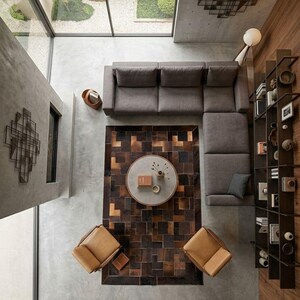 Op de afbeelding: Een bovenaanzicht van een moderne woonkamer met een grijze hoekbank, een ronde salontafel en twee bruinleren stoelen. Een patchwork tapijt in bruin- en zwarttinten ligt onder de tafel. Een groot raam biedt uitzicht op een tuin.