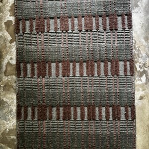 May include: A rectangular woven rug featuring a repeating geometric pattern of dark charcoal grey and deep brown vertical bars. The texture consists of alternating horizontal ribbed sections and raised, block-like vertical segments.