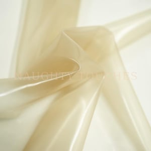 Può includere: Un foglio di materiale in lattice traslucido color beige pallido con una finitura liscia e lucida. Il materiale è drappeggiato per creare pieghe e curve morbide. Il testo NAUGHTY TOUCHES è stampato in un carattere chiaro e sottile.