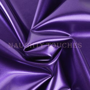 May include: A piece of deep purple fabric with a smooth, reflective, and metallic finish. The material is draped to create soft folds and shadows. The text NAUGHTY TOUCHES is printed in a light, neutral colour across the centre of the fabric.