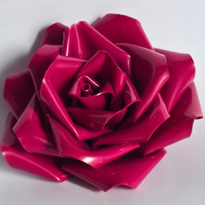 May include: A close-up of a vibrant, magenta-coloured rose. The petals are tightly wound in the centre, gradually unfurling outwards. The glossy surface reflects light, highlighting the intricate folds and curves of the flower. The rose is set against a plain white background.