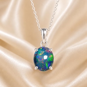May include: A silver necklace featuring a round, iridescent opal pendant. The opal displays a mix of blue, green, and red hues. The pendant is held by four prongs and hangs from a delicate silver chain. The background is a soft, satin fabric.