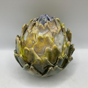 May include: A ceramic artichoke sculpture with a detailed, textured surface. The artichoke features a blend of colours, including green, yellow, and blue, creating a naturalistic appearance. The sculpture is a decorative item.