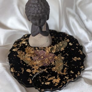 Puede incluir: Una pequeña estatua de Buda gris oscuro con las manos juntas en oración, sobre un posavasos negro de forma irregular. El posavasos está decorado con detalles de pan de oro y un toque de rosa. La estatua mide unos 7.6 cm de altura.