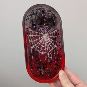 Op de afbeelding: Ovale harsbak met een Halloween-thema. De bak heeft een rood-zwarte gradiënt achtergrond met een wit spinnenwebontwerp en zwarte vleermuisvormige glitter. De bak wordt door een hand vastgehouden.