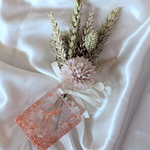 Puede incluir: Un prendido hecho a mano con trigo seco, una flor rosa pálido y un rectángulo de resina transparente con escamas de cobre. El prendido está atado con una cinta blanca, sobre un fondo de satén blanco.
