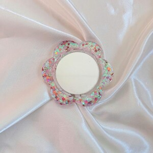 Può includere: Un supporto per telefono a forma di fiore con una cornice in resina trasparente incastonata con glitter iridescenti multicolori e piccoli accenti a forma di cuore. Il centro dell'accessorio contiene una superficie a specchio circolare riflettente.