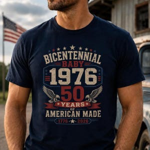 May include: A navy blue short-sleeve t-shirt featuring a graphic print with the text BICENTENNIAL BABY 1976 50 YEARS OF AMERICAN MADE 1776-2026. The design includes two eagles, stars, and red, white, and blue accents in a distressed vintage style.