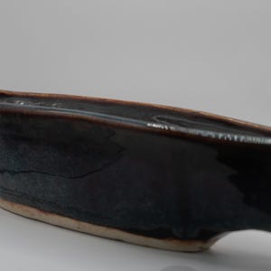 Pode incluir: Um prato de cerâmica azul escuro feito à mão, com formato retangular e uma borda ligeiramente curvada. O prato tem um acabamento brilhante e uma base bege clara. O interior é um tom de azul mais escuro.