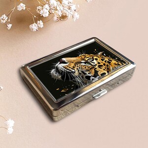 May include: A rectangular, silver-toned metal cigarette case featuring a detailed illustration of a jaguar's head against a black background. The jaguar has a tan and black spotted coat, green eyes, and white whiskers. The case is closed with a small clasp.