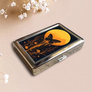 以下が含まれることがあります： 黒とオレンジ色のグラフィックが施されたシルバーの金属製シガレットケース。グラフィックは、オレンジ色の枝と花のある、黄色の月の前にいる蝶を描いています。ケースは長方形で、ヒンジ付きの蓋が付いています。