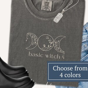 May include: A charcoal gray crewneck t-shirt featuring a white line art graphic of a triple moon phase symbol with floral details and the text basic witch. The shirt is paired with blue denim jeans, black leather boots, and a silver pendant necklace.