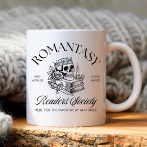 May include: White ceramic mug with the word "ROMANTASY" in black script. The mug features a skull wearing a crown, a rose, a candle, and a dagger. Text on the mug reads "Readers Society" and "Here for the swordplay and spice."